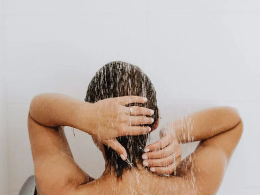 Female rinsing hair under a shower with water streaming down their back in a tiled bathroom.